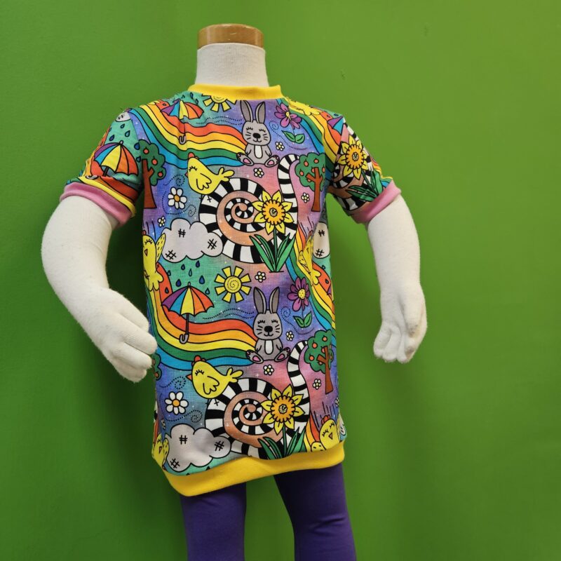 A child mannequin wears a tshirt dress made from Spring Adventures bright and fun jersey with yellow trim and purple leggings underneath