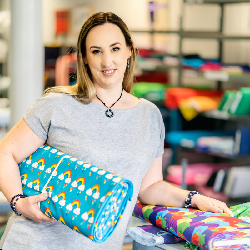 A lady holds several bolts of brightly coloured fabrics and smiles at the camera.