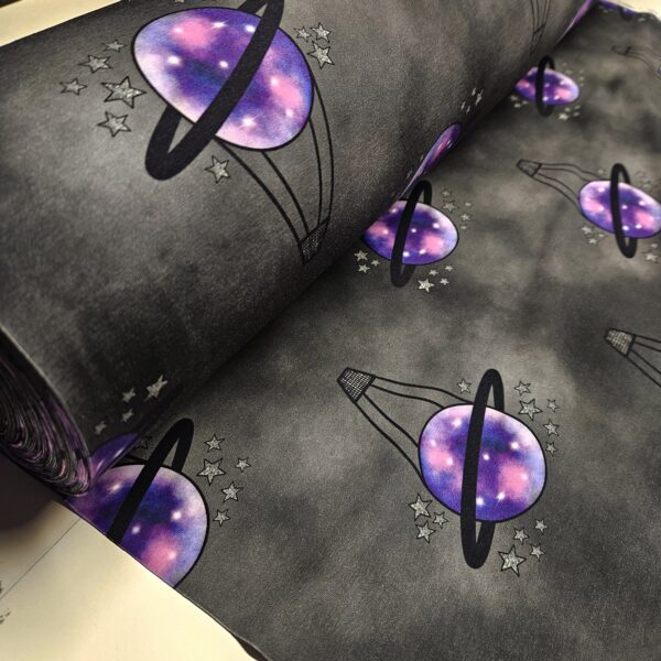 A black and grey mottled fabric with a hot air balloon repeating motif. The hot air balloon has a small black basket and a purple planet balloon with black saturn ring around it. Small silver stars surround the balloon.