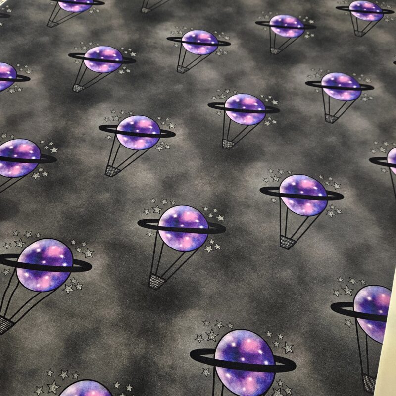 A black and grey mottled fabric with a hot air balloon repeating motif. The hot air balloon has a small black basket and a purple planet balloon with black saturn ring around it. Small silver stars surround the balloon.