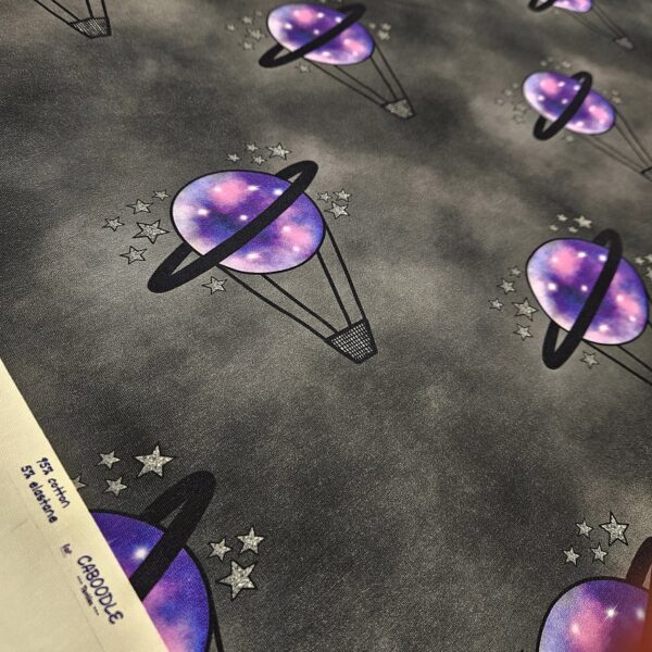 A black and grey mottled fabric with a hot air balloon repeating motif. The hot air balloon has a small black basket and a purple planet balloon with black saturn ring around it. Small silver stars surround the balloon.