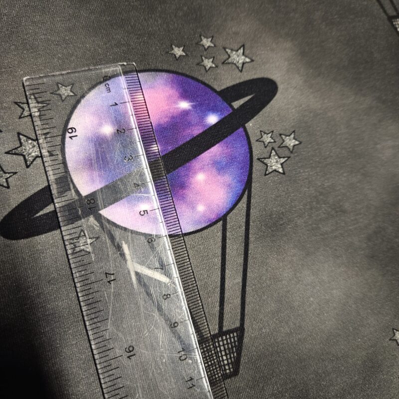 A black and grey mottled fabric with a hot air balloon repeating motif. The hot air balloon has a small black basket and a purple planet balloon with black saturn ring around it. Small silver stars surround the balloon. A ruler shows that the circle of the balloon measures 6cm and the total height of the balloon is 11cm