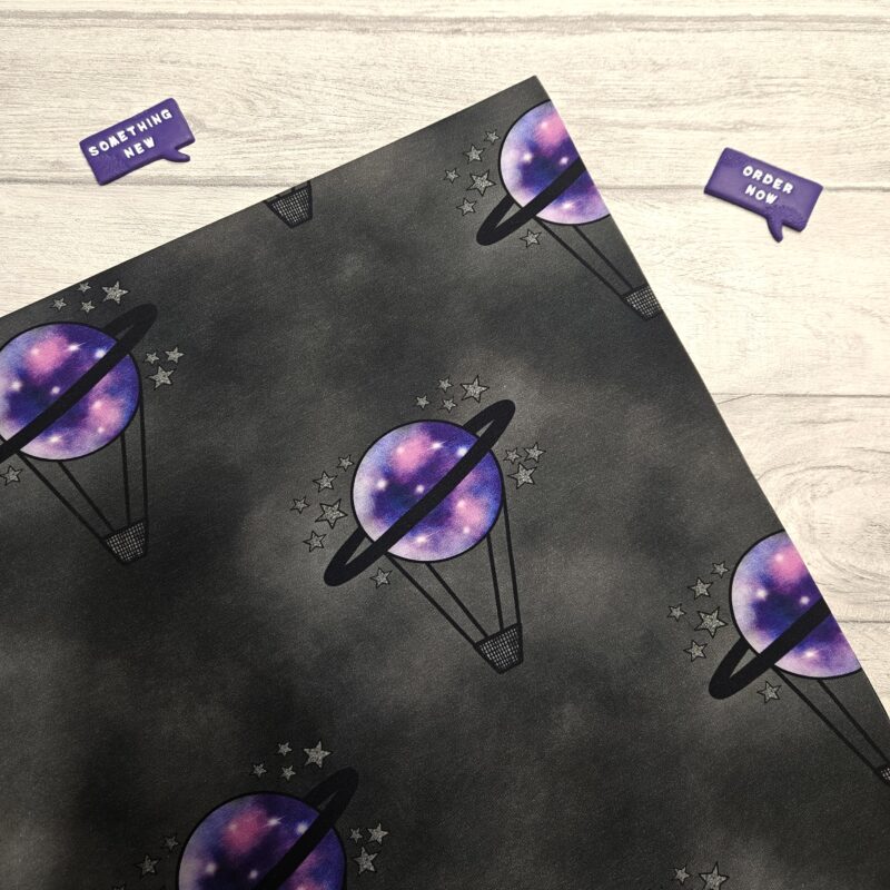 A black and grey mottled fabric with a hot air balloon repeating motif. The hot air balloon has a small black basket and a purple planet balloon with black saturn ring around it. Small silver stars surround the balloon.