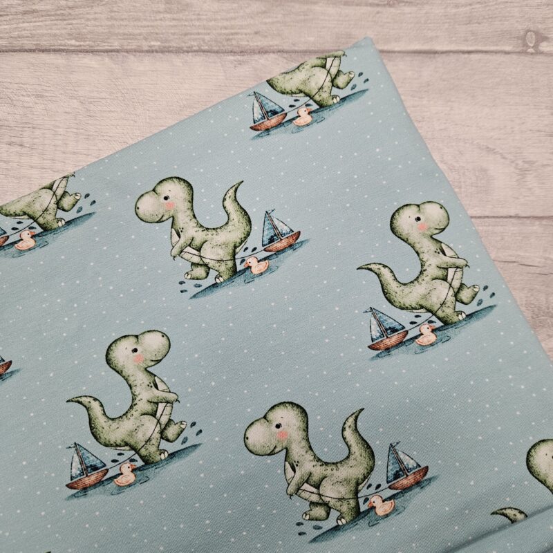 Dino's Bath Time Jersey Fabric is a fabric with a pale blue back ground and dino pulling along his little boat through splashes. He is accompanied by his rubber duck