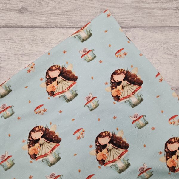 Mushroom Fairy Jersey Fabric is a light blue background fabric with a long haired fairy sitting on top of a red spotted toadstool mushroom. She carries a squirrel on her knee